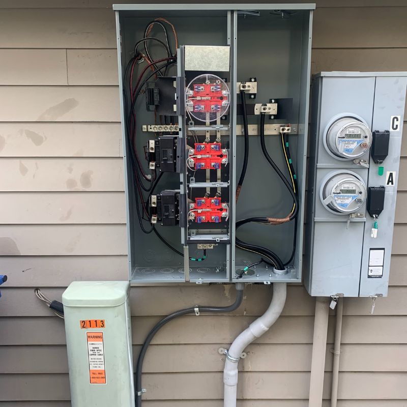 Open outdoor electrical panel showing meters, wiring, and circuit breakers on a building exterior.