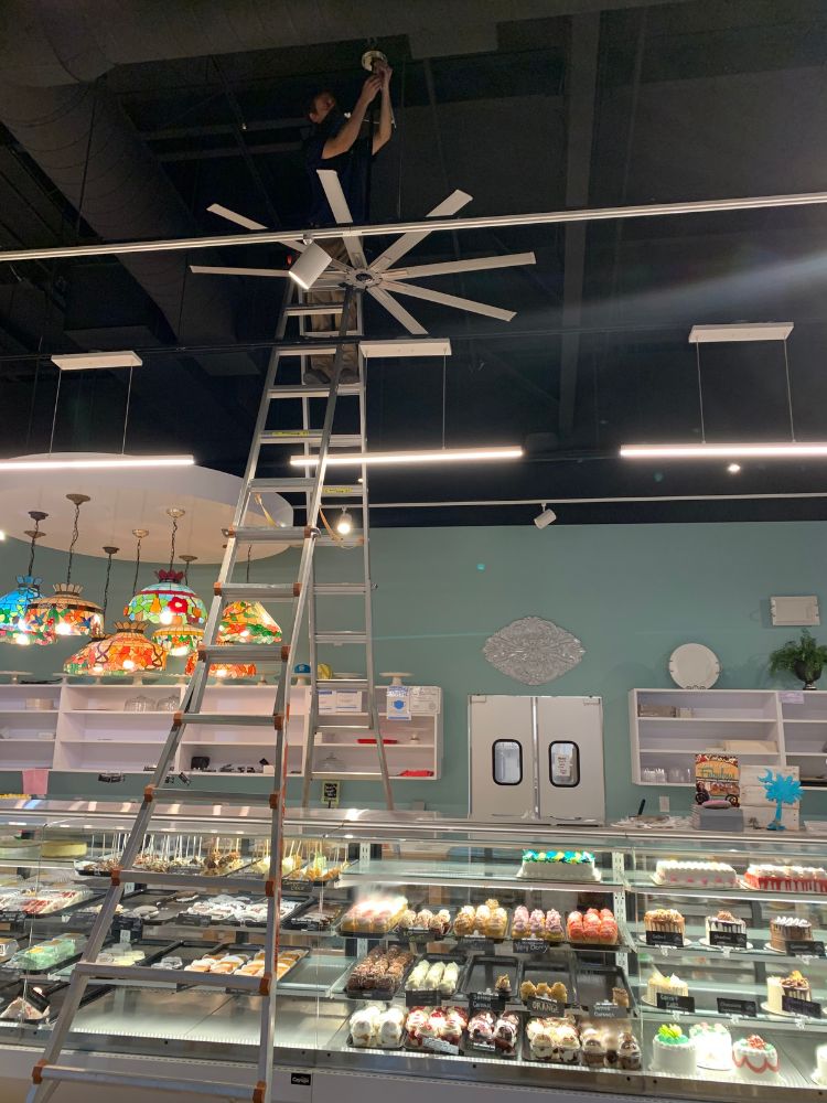 Electrician using a power drill to mount device in ceiling tile with overhead lighting and exposed framing.