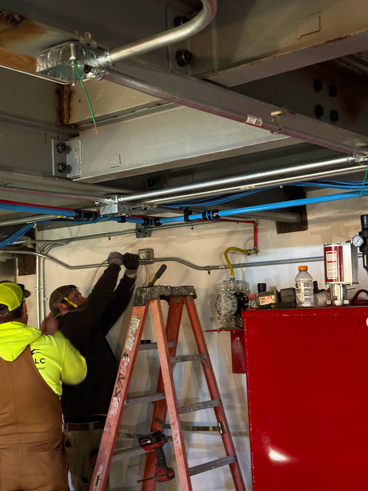 Electrician using a power drill to mount device in ceiling tile with overhead lighting and exposed framing.