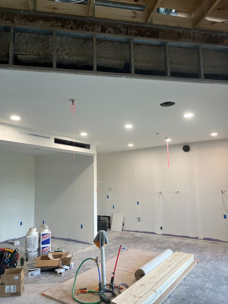 Electrician using a power drill to mount device in ceiling tile with overhead lighting and exposed framing.